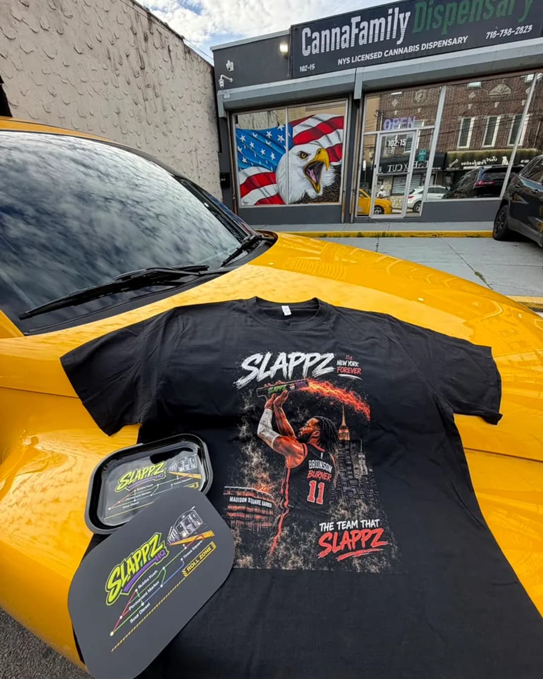 A SLAPPZ t-shirt and tin laid out on the hood of a yellow cab outside the CannaFamily Dispensary storefront.