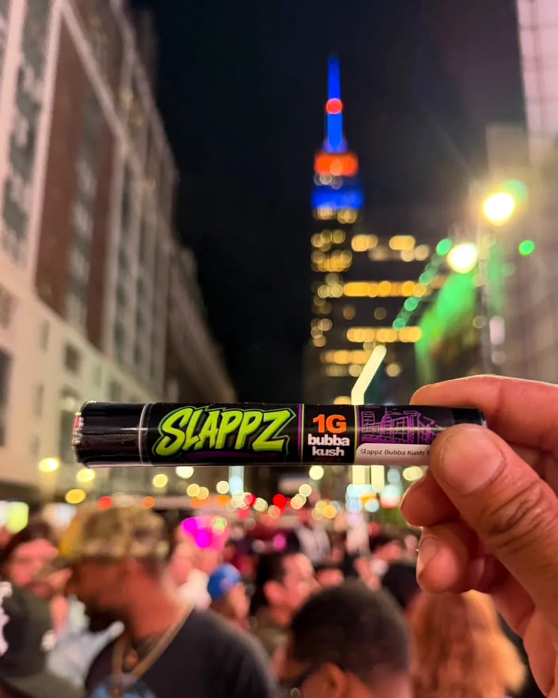 A SLAPPZ 1g Bubba Kush pre-roll tube held up over a packed night-time street, a floodlit tower behind it.