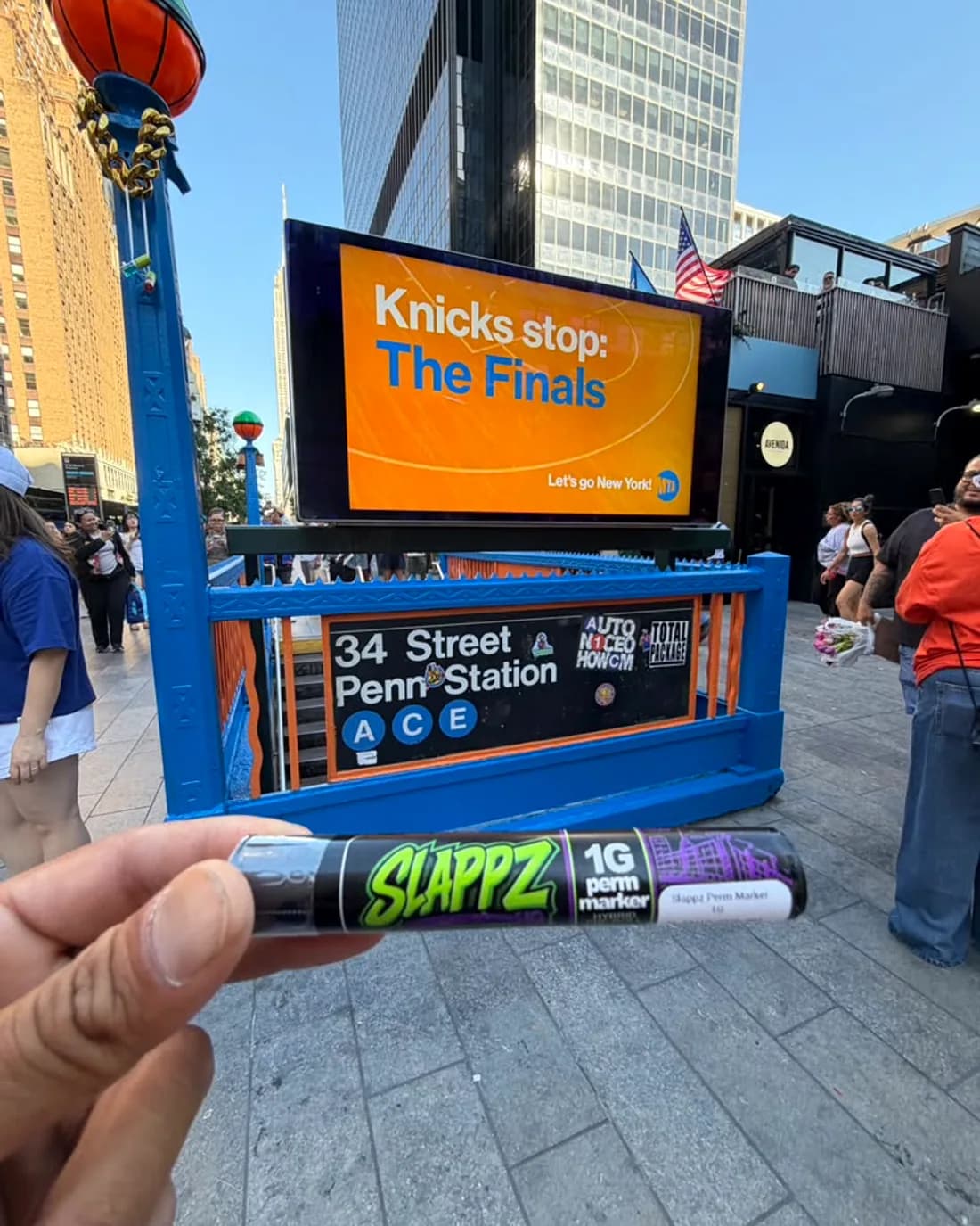 A SLAPPZ 1g Perm Marker pre-roll tube held up in front of the 34 Street–Penn Station A, C, E subway entrance.