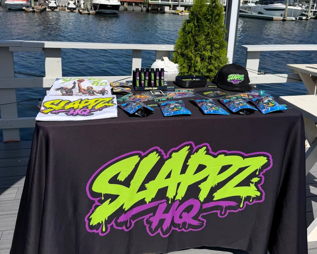 A SLAPPZ HQ branded table set up on a waterfront deck, product and caps laid out, boats moored behind.